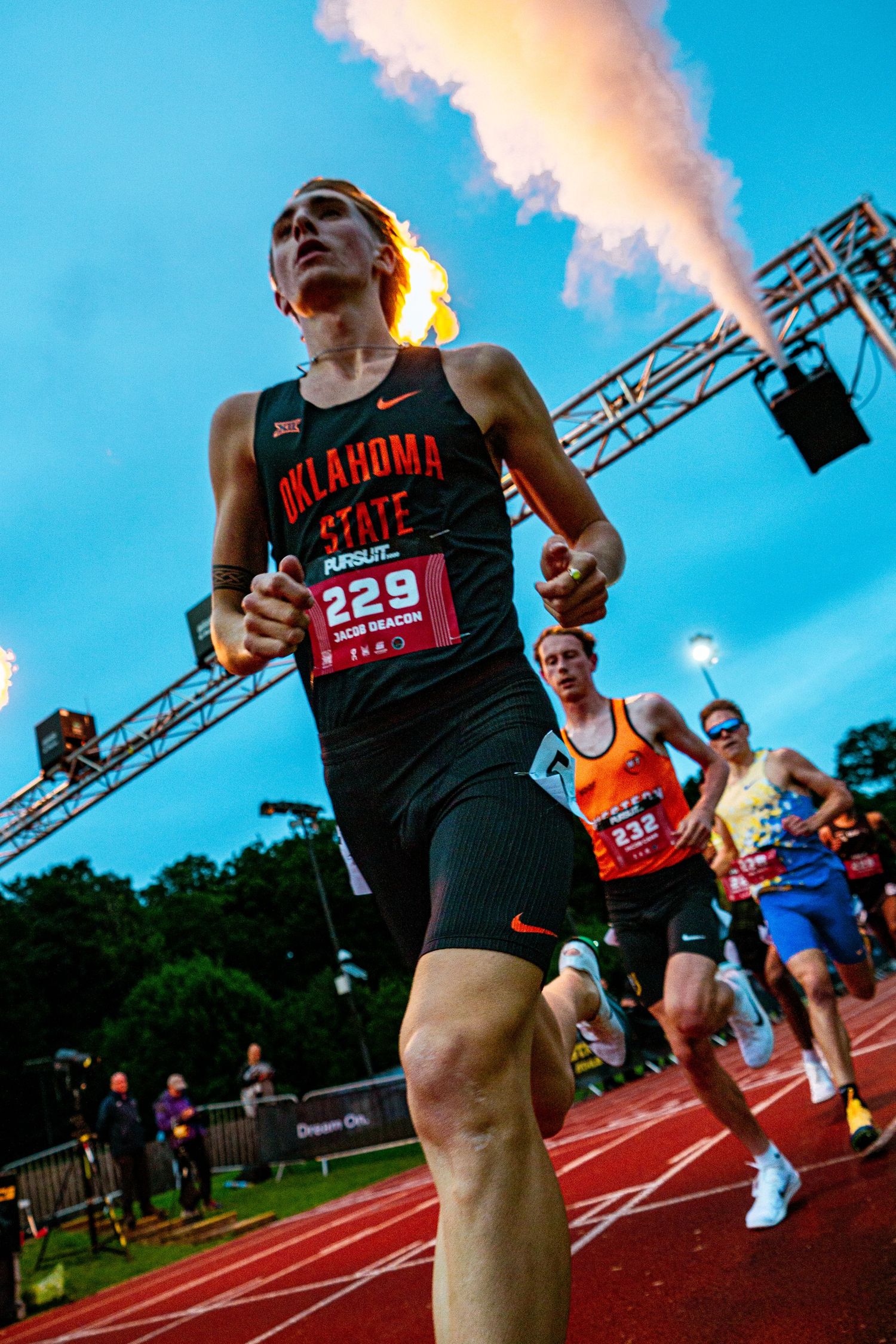 Oklahoma State runner #229 sprints past competitors on a track with colorful smoke effects at a night racing event.