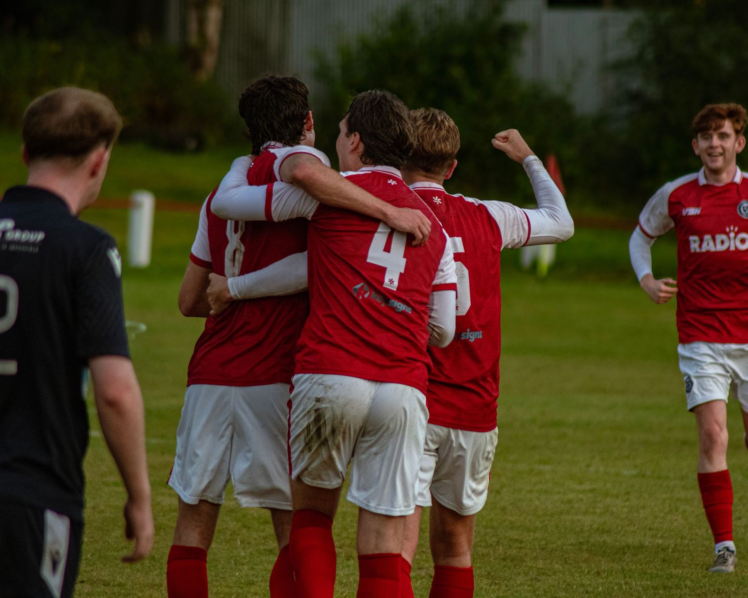Soccer players in red and white Radio jerseys celebrating a goal on a green field.