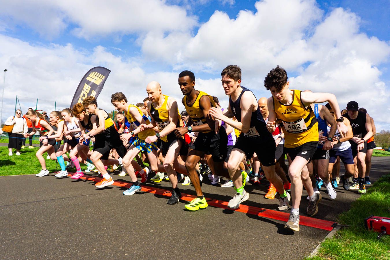 Large group of male runners in yellow vests sprint from the start line at an outdoor athletics race.
