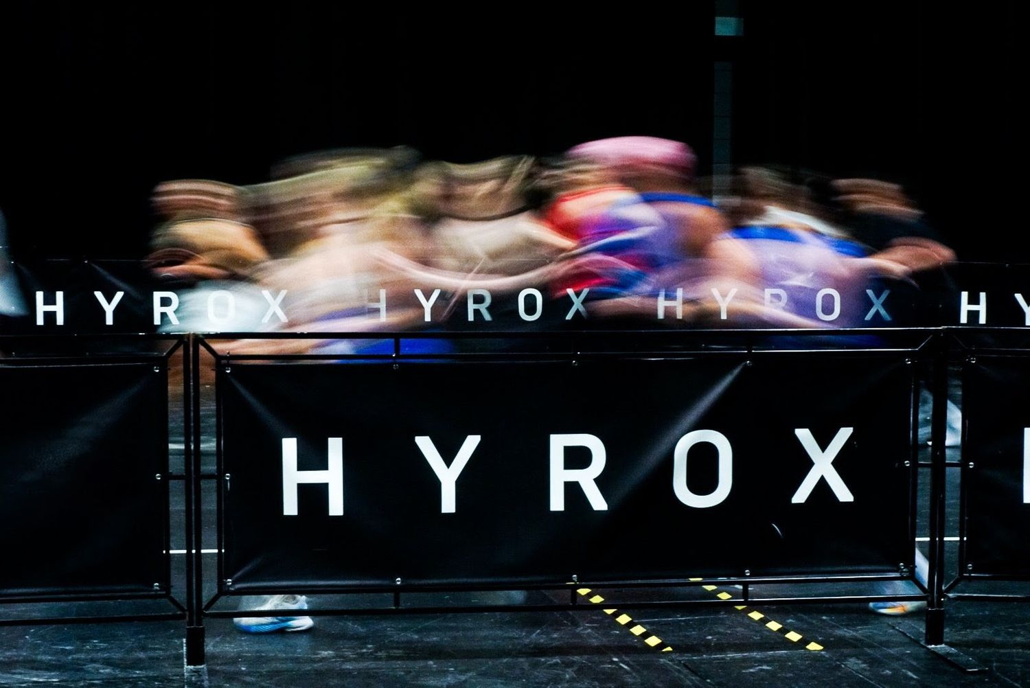 Athletes compete in a HYROX fitness race event under dramatic lighting with motion blur effect.