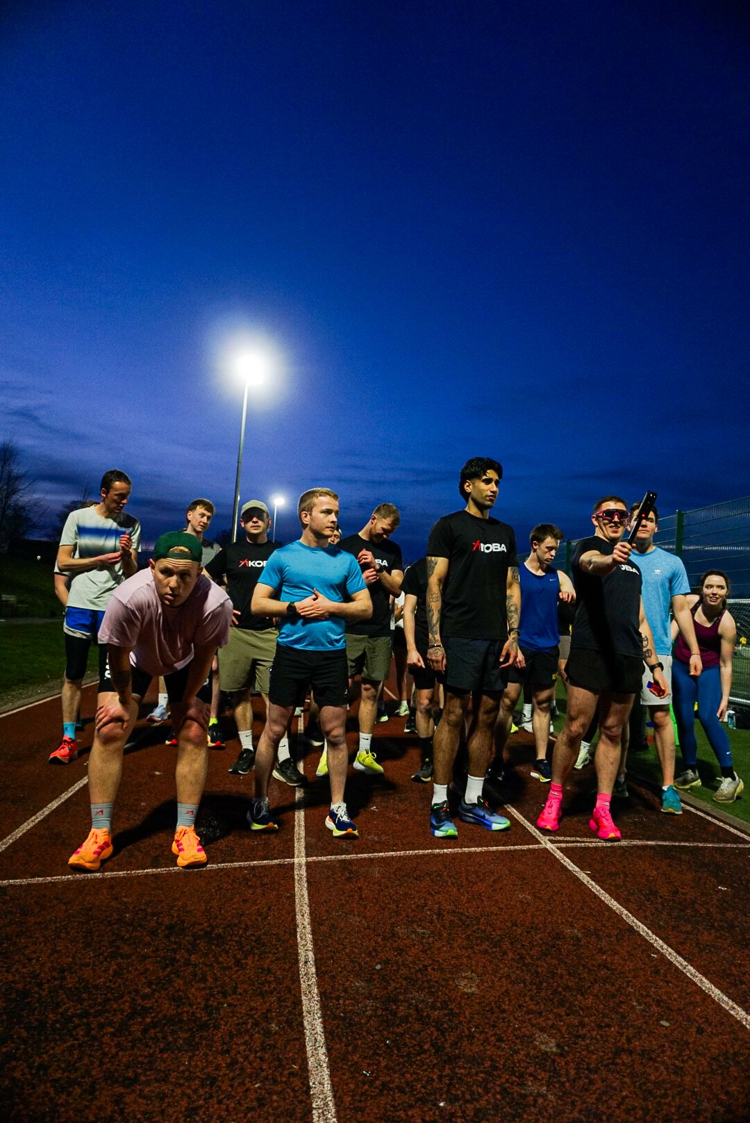 Large group of runners preparing to race on a red athletic track under floodlights at dusk.