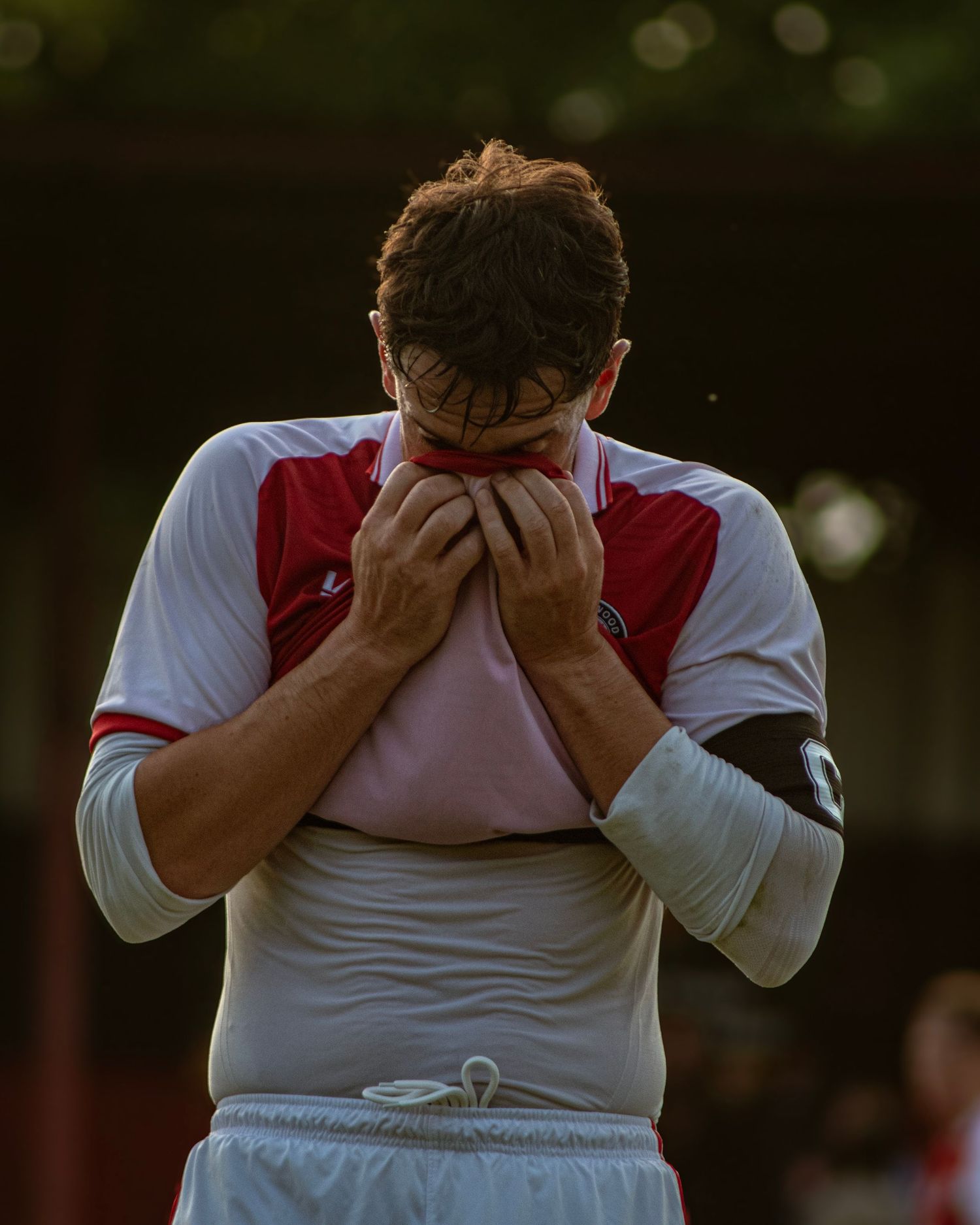 A footballer in red and white kit covers his face with his jersey in frustration.