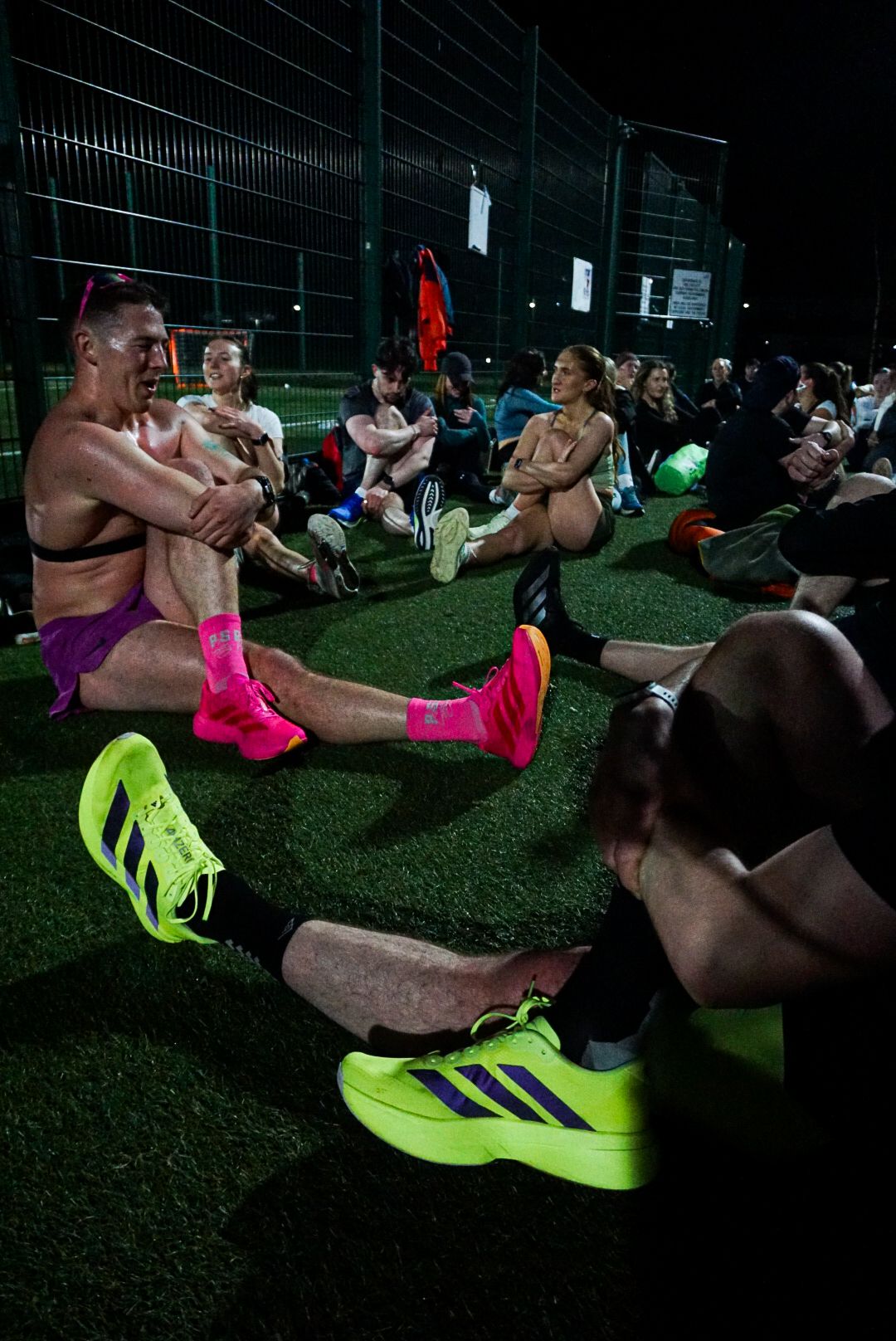 Athletes in colorful neon running shoes resting on artificial grass after a night track session.