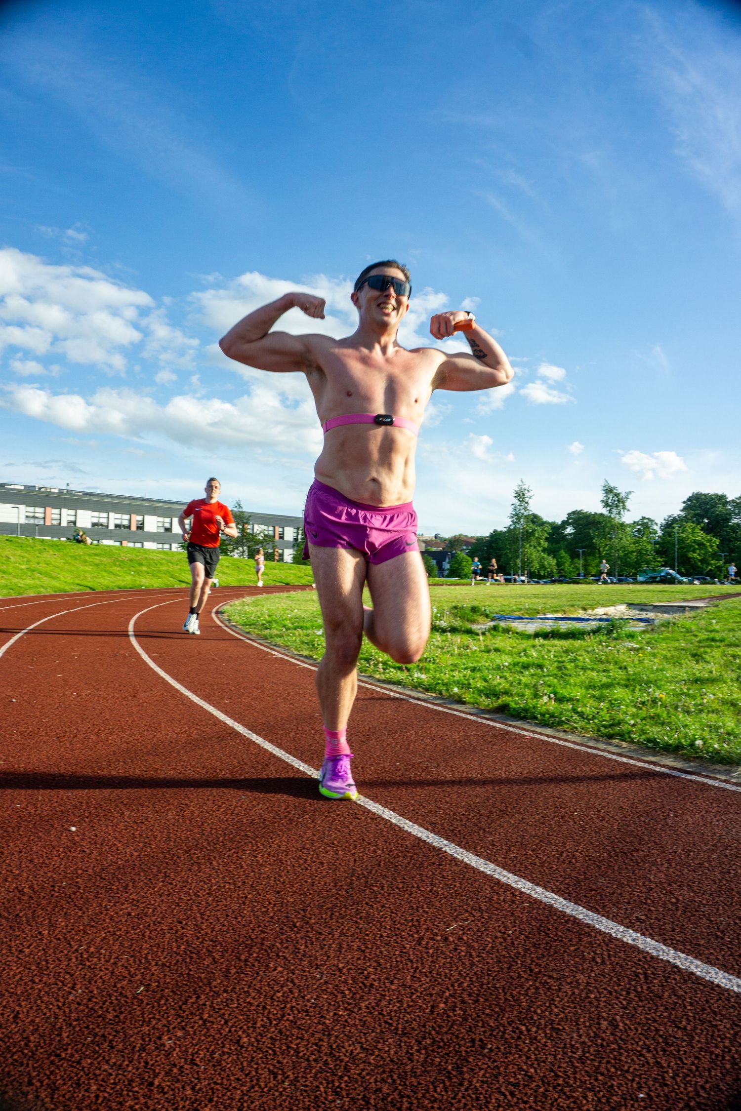 Shirtless male runner in pink shorts flexing muscles while running on outdoor athletics track.