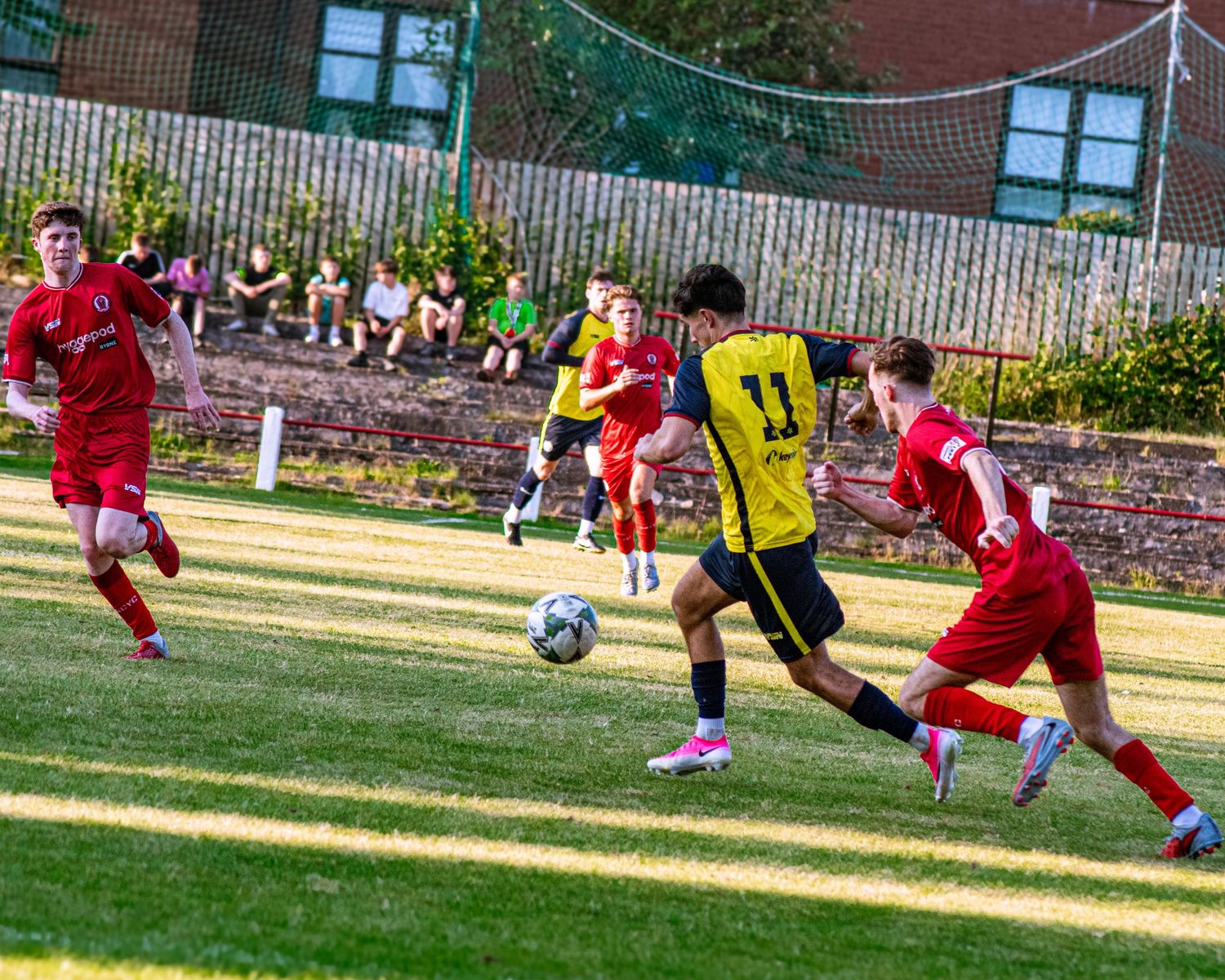Player in yellow number 11 jersey dribbles soccer ball past red-uniformed defenders on green pitch.