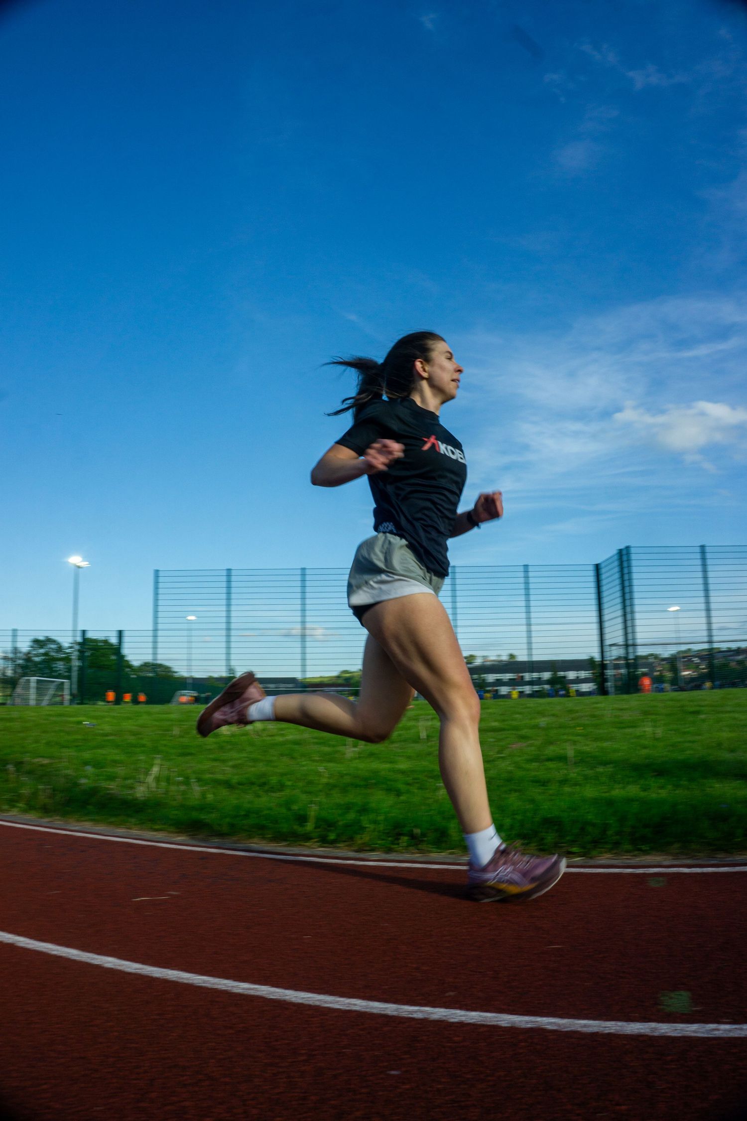 Female athlete in black top running on red track against a vivid blue sky.