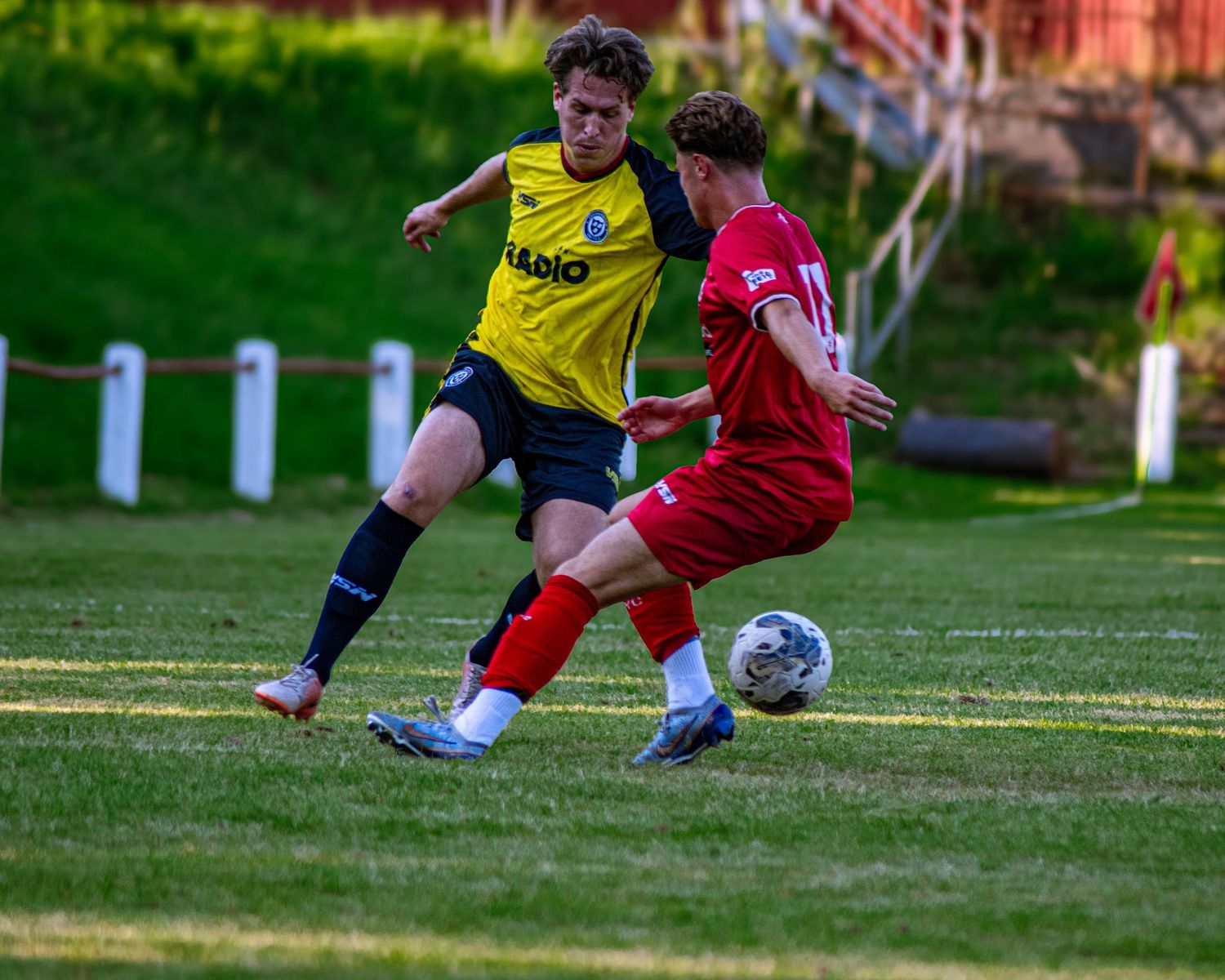 Two soccer players compete for the ball, one in yellow and one in red, during a match on a grass field.