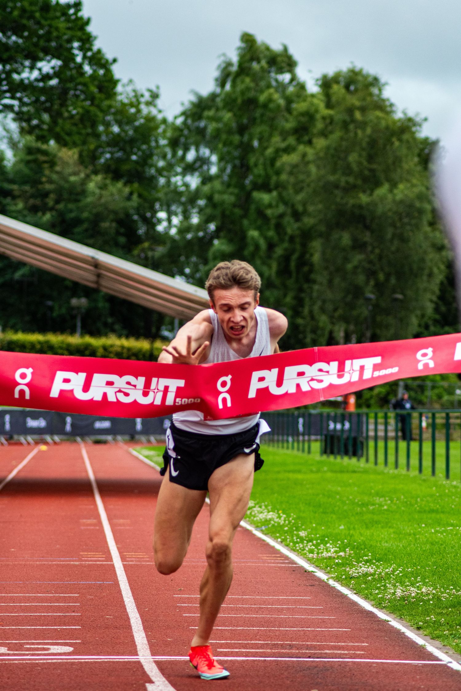 Male runner breaks through the finish line tape at the On Pursuit track event on a red athletics track.
