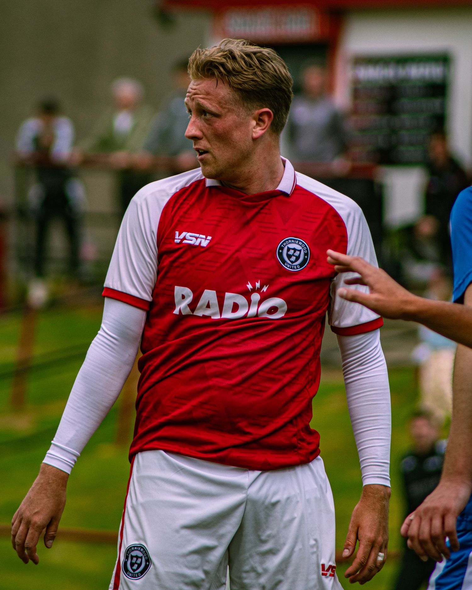 Male soccer player in red and white VSN Radio jersey and white shorts standing on a football pitch.