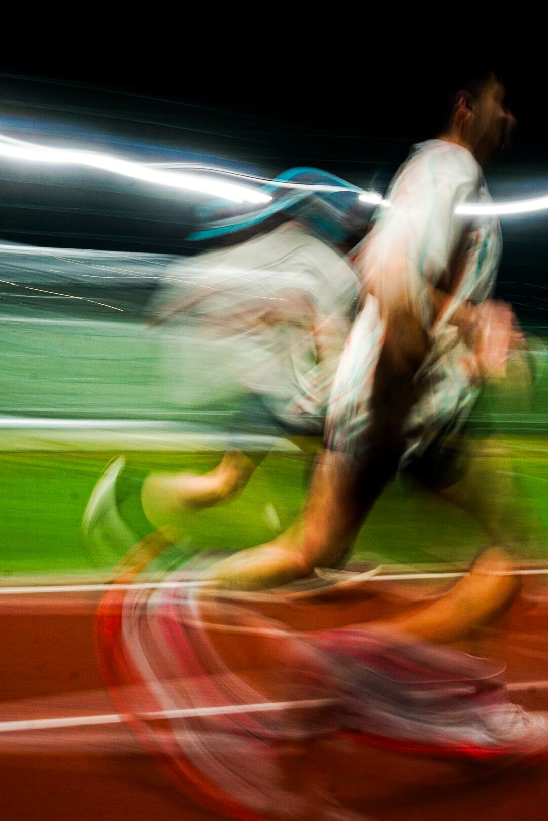 Dynamic motion blur photograph of a sprinter racing on a red track under stadium lights at night.