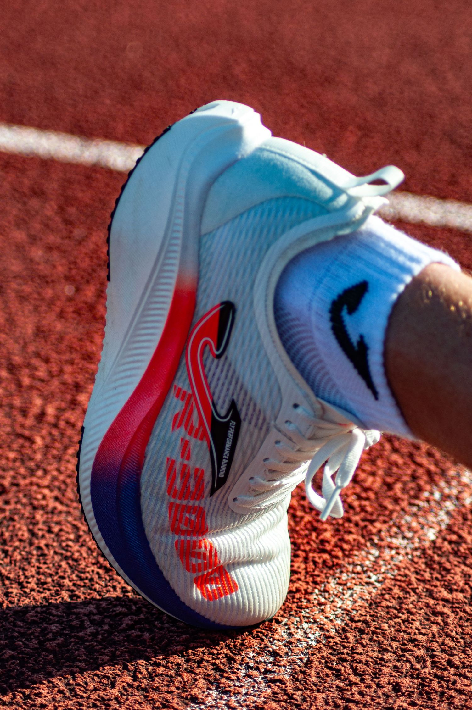 Close-up of a white and red performance running shoe on a red athletics track surface in bright sunlight.