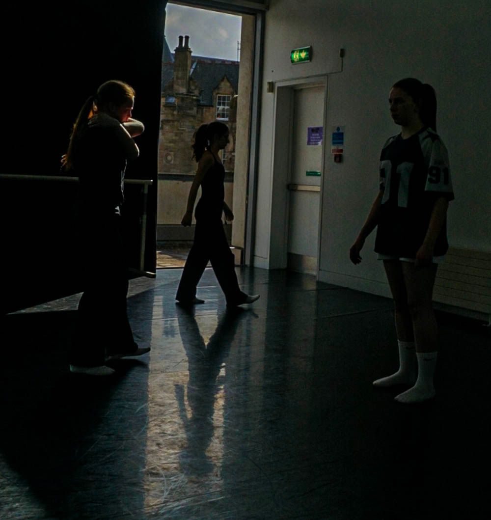 Three silhouetted figures stand in a dimly lit dance studio with light streaming through a window.