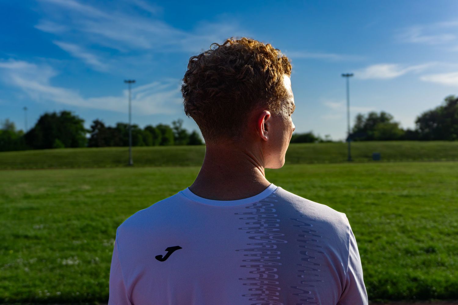 Rear view of a young curly-haired male athlete in a white Joma shirt looking out over a green sports field.