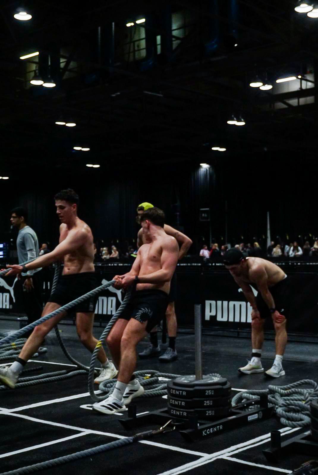 Shirtless male athletes pushing a weighted sled with battle ropes at a PUMA fitness competition.