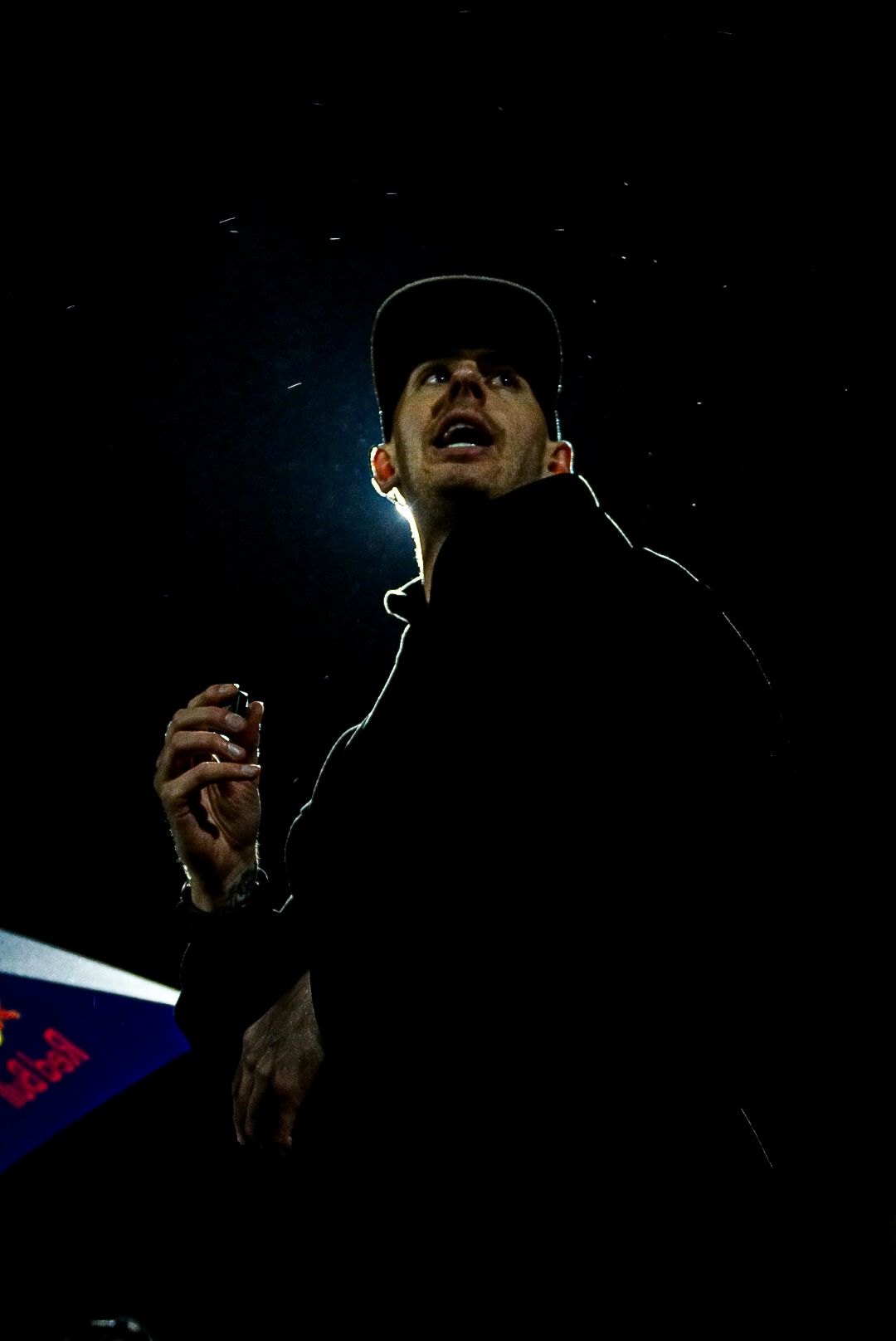 Silhouetted man wearing a cap looking upward dramatically lit from behind in darkness.