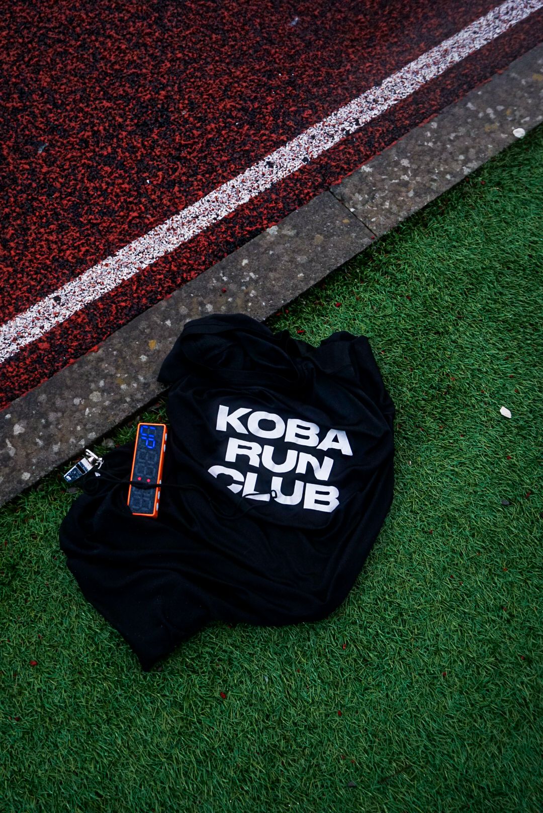 Black Koba Run Club t-shirt lying on green grass beside red running track.