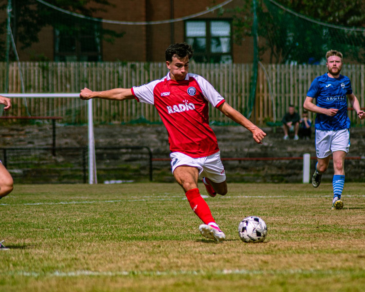 Soccer player in red and white kit dribbling ball on grass pitch during competitive match.