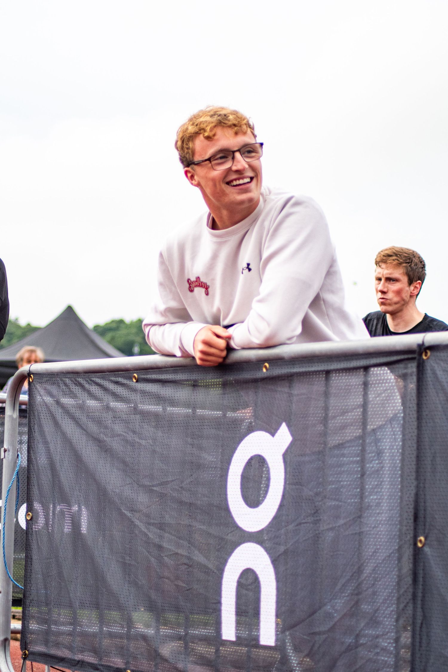 Smiling young man with glasses leaning on a metal barrier at an outdoor festival event.