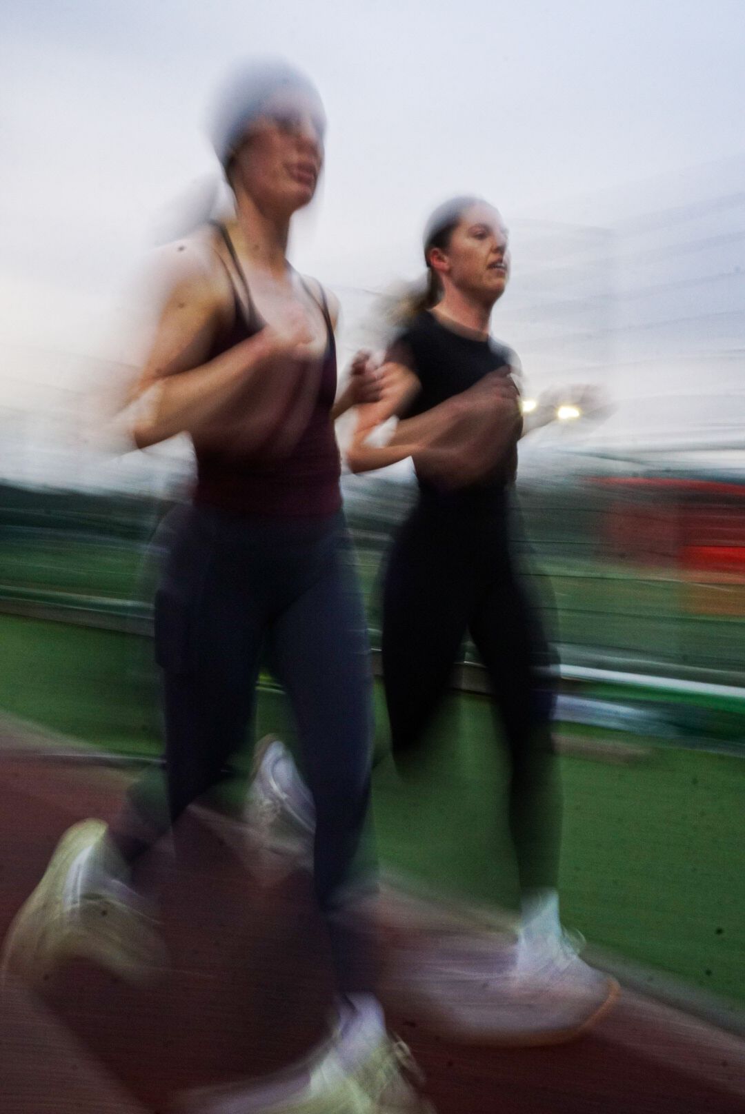 Two female runners in motion blur on red athletics track at dusk.