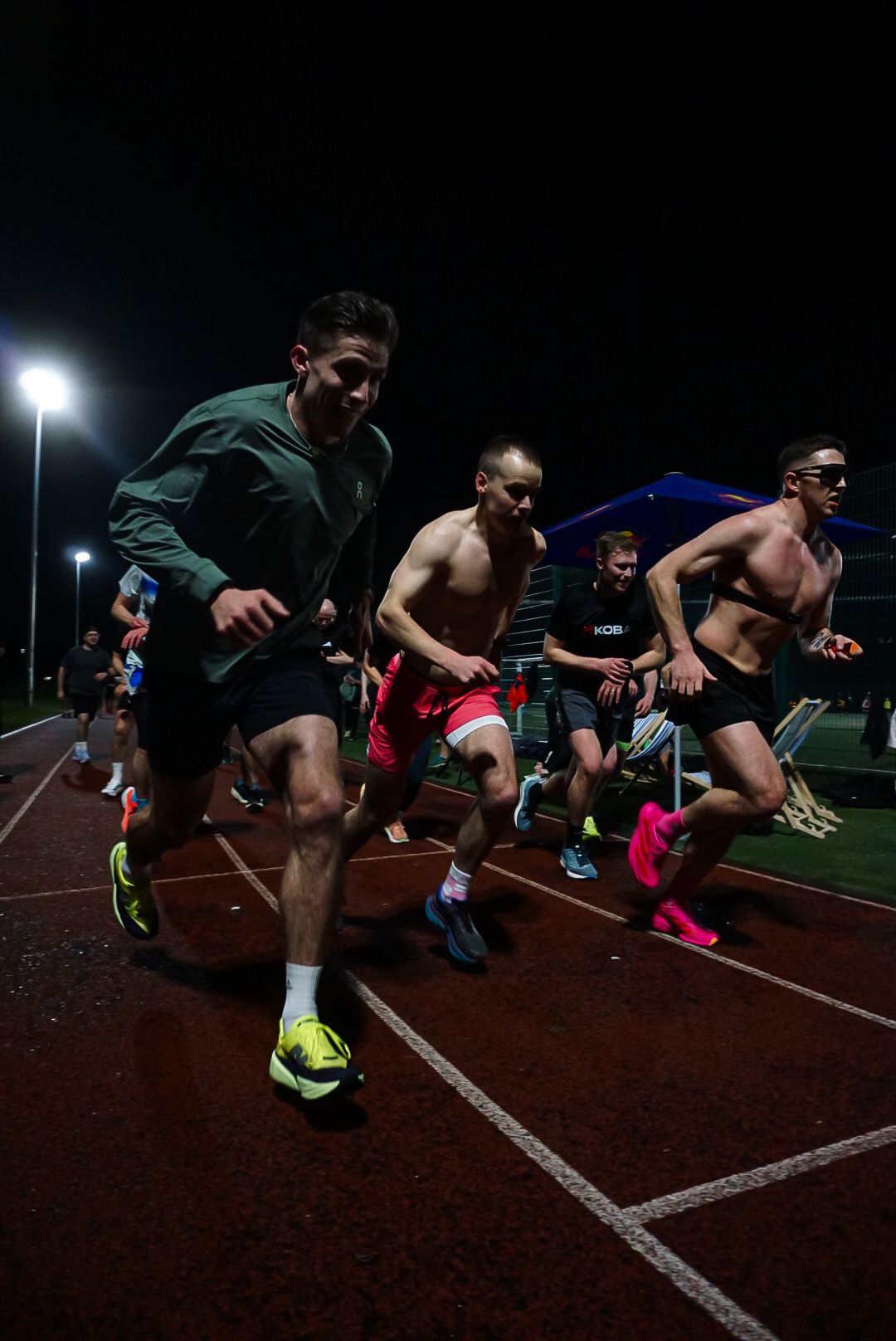 Group of male runners racing on an outdoor track at night under bright floodlights.