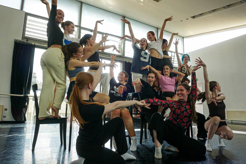 A large group of energetic dance students strike expressive poses together in a bright studio with ballet barres.