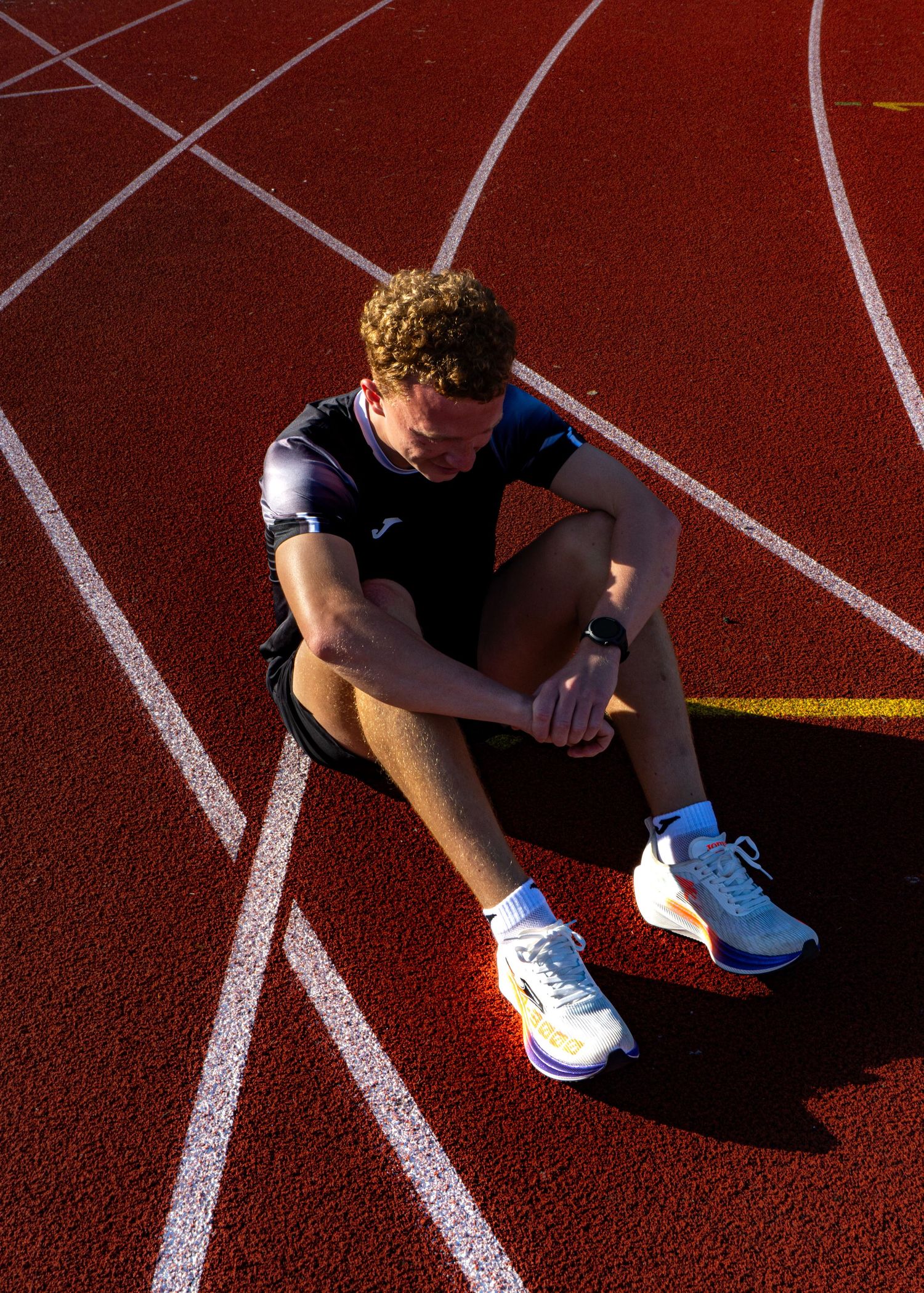 Male track athlete sitting on red running track lanes, wearing Joma kit and white performance running shoes.