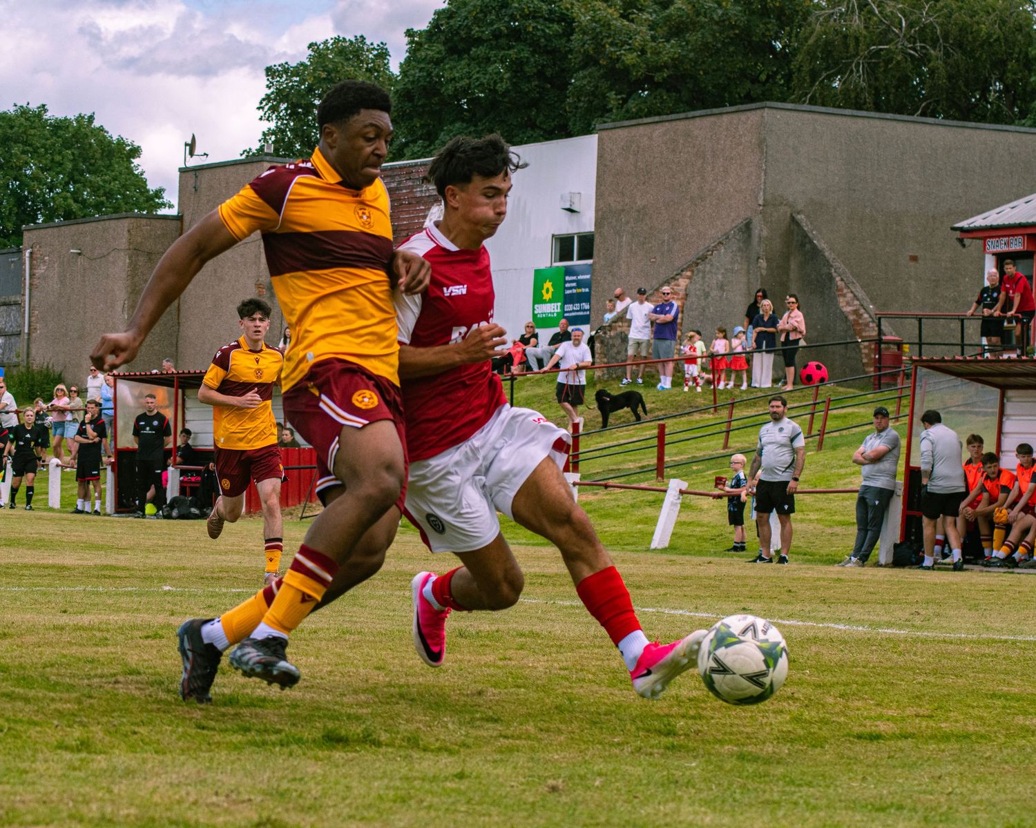 Two soccer players compete for the ball during a match on a grass field with spectators watching in the background.
