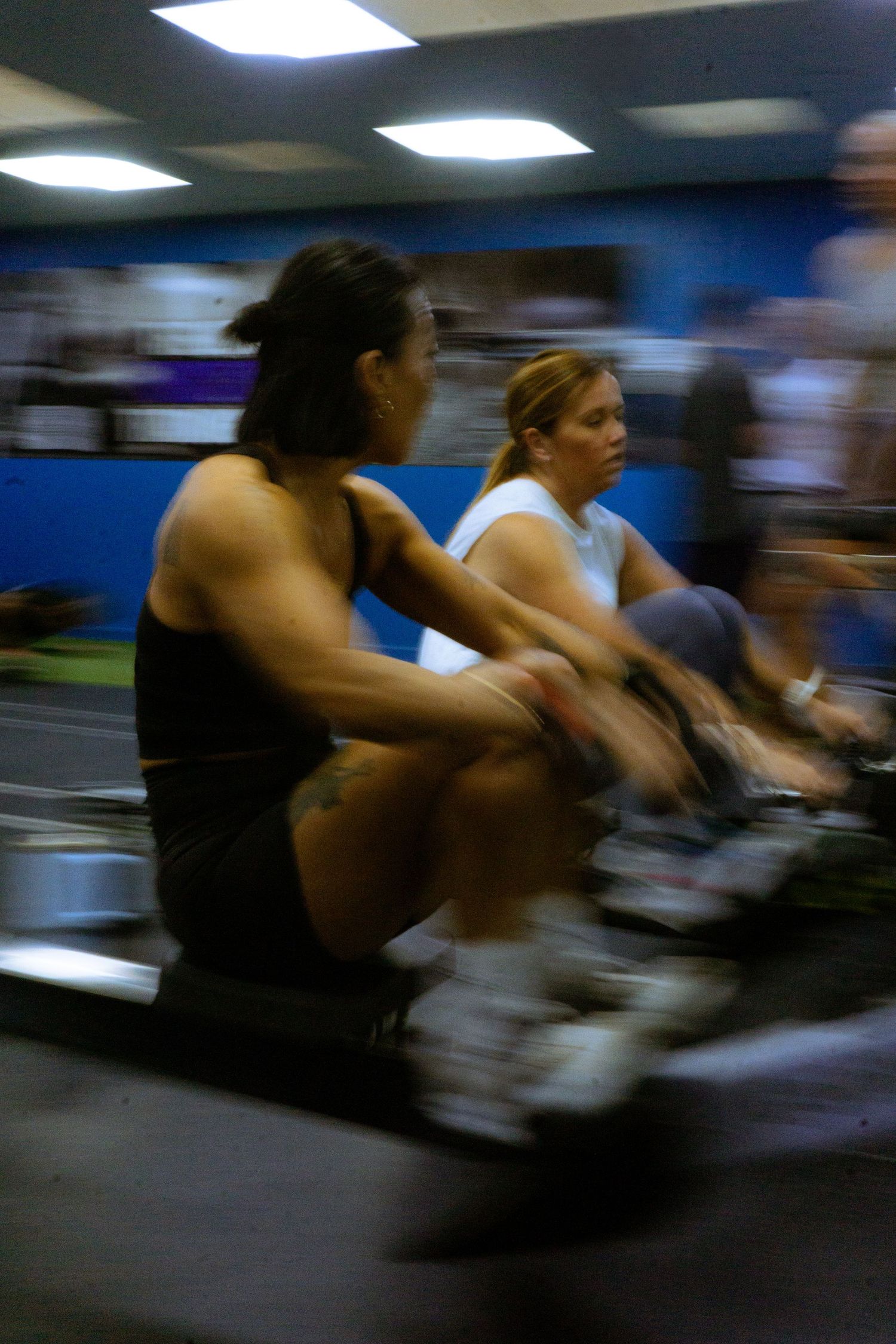 Two women exercising on rowing machines in a gym, captured with motion blur effect.