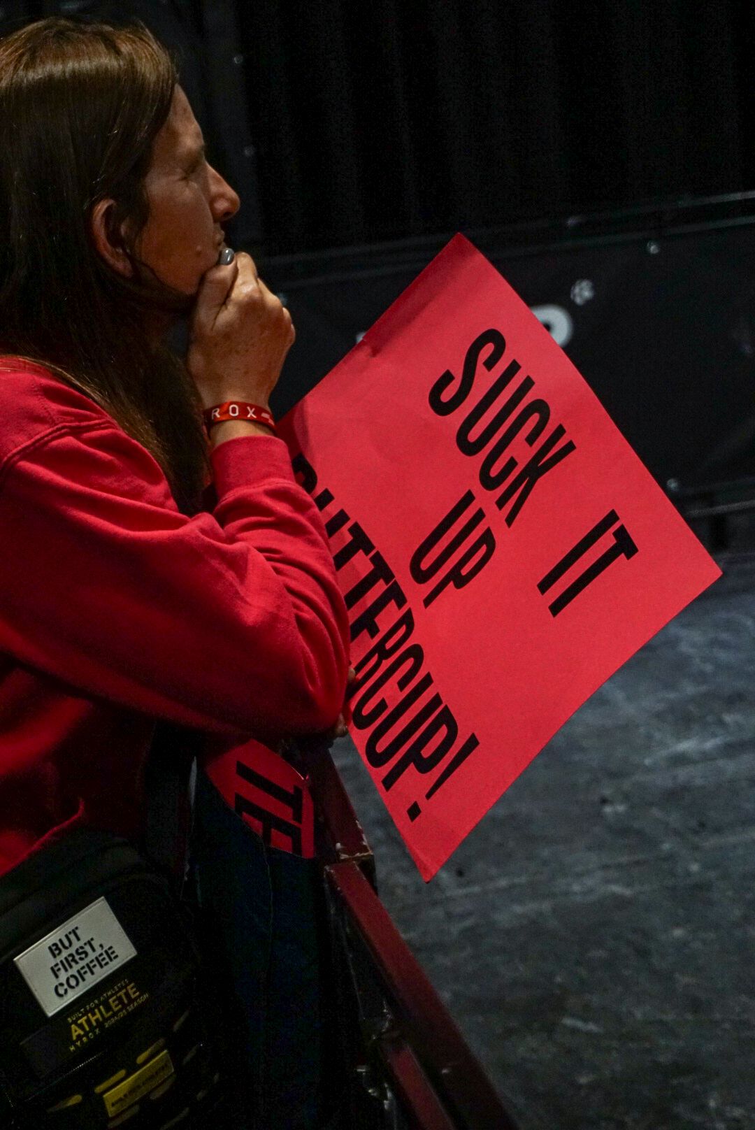 Spectator in red hoodie holds a Suck It Up Buttercup motivational sign at a HYROX event.