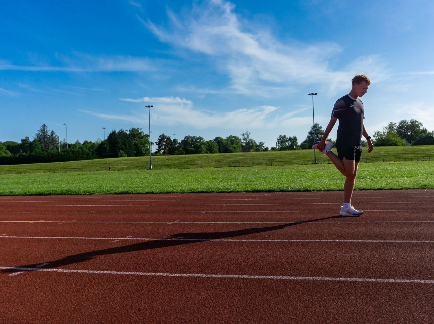 Male athlete stretching on a red athletics track with green fields and blue sky behind.