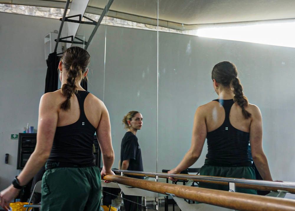 Two female ballet dancers in black practice wear stand at barre facing mirror in dance studio.