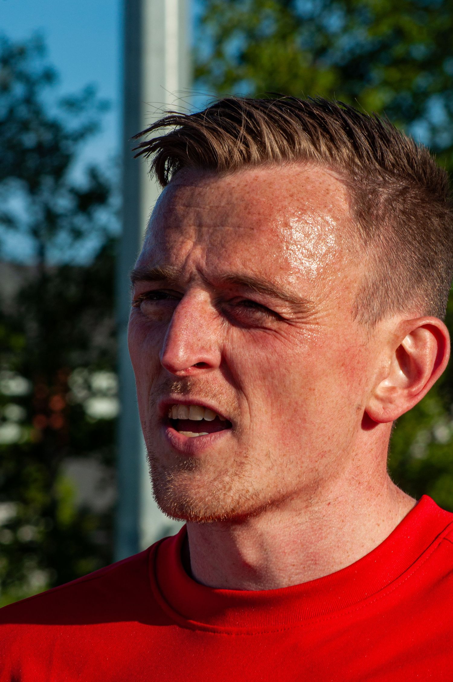 Sweaty male athlete in red shirt with slicked-back hair at an outdoor running event.