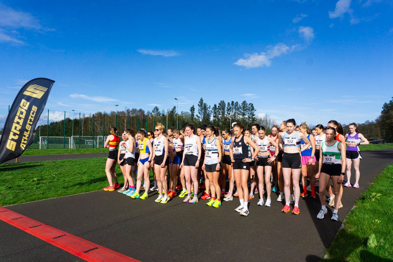 Female runners line up at the start line of an outdoor road race on a sunny day.