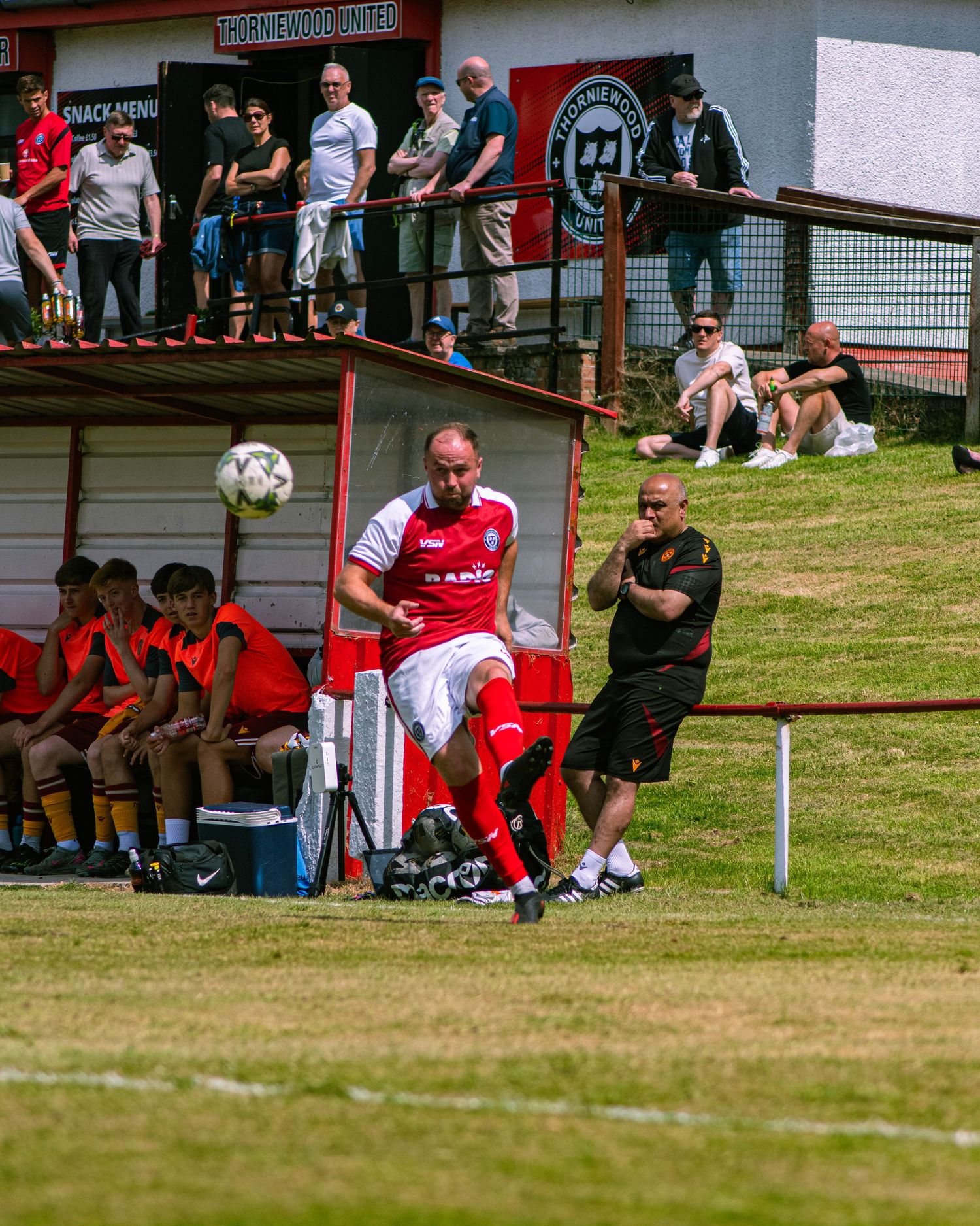 A soccer player in red and white kicks the ball near the dugout during a sunny match at Thornewood United FC.