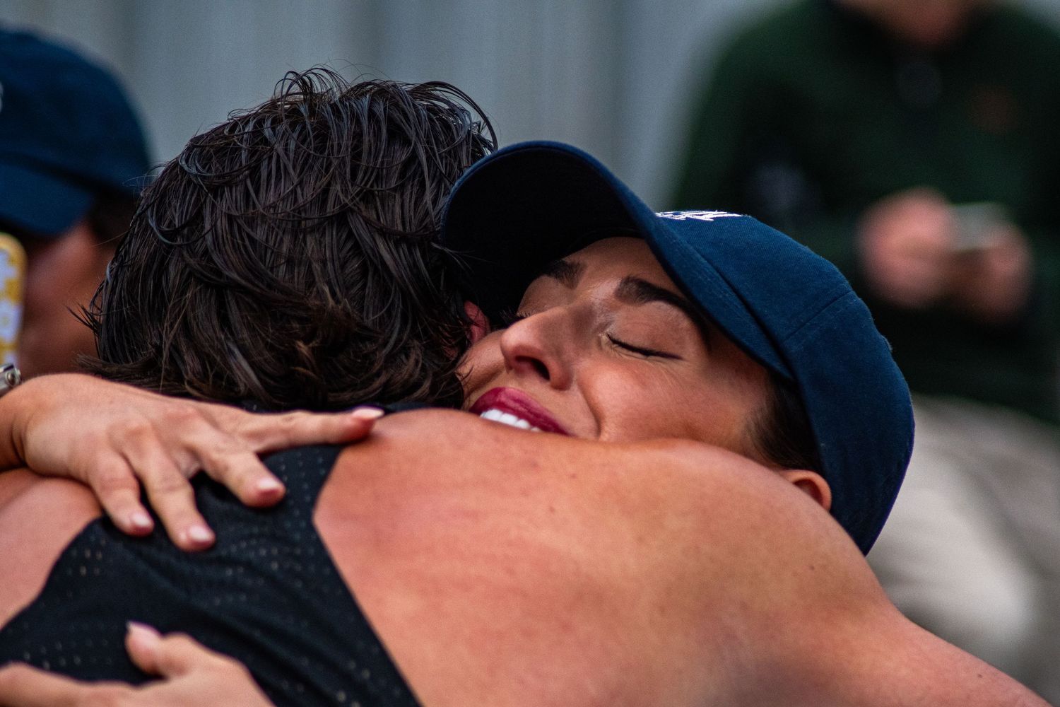 Two people sharing an emotional hug, with a woman in a blue cap smiling with eyes closed in a heartfelt embrace.