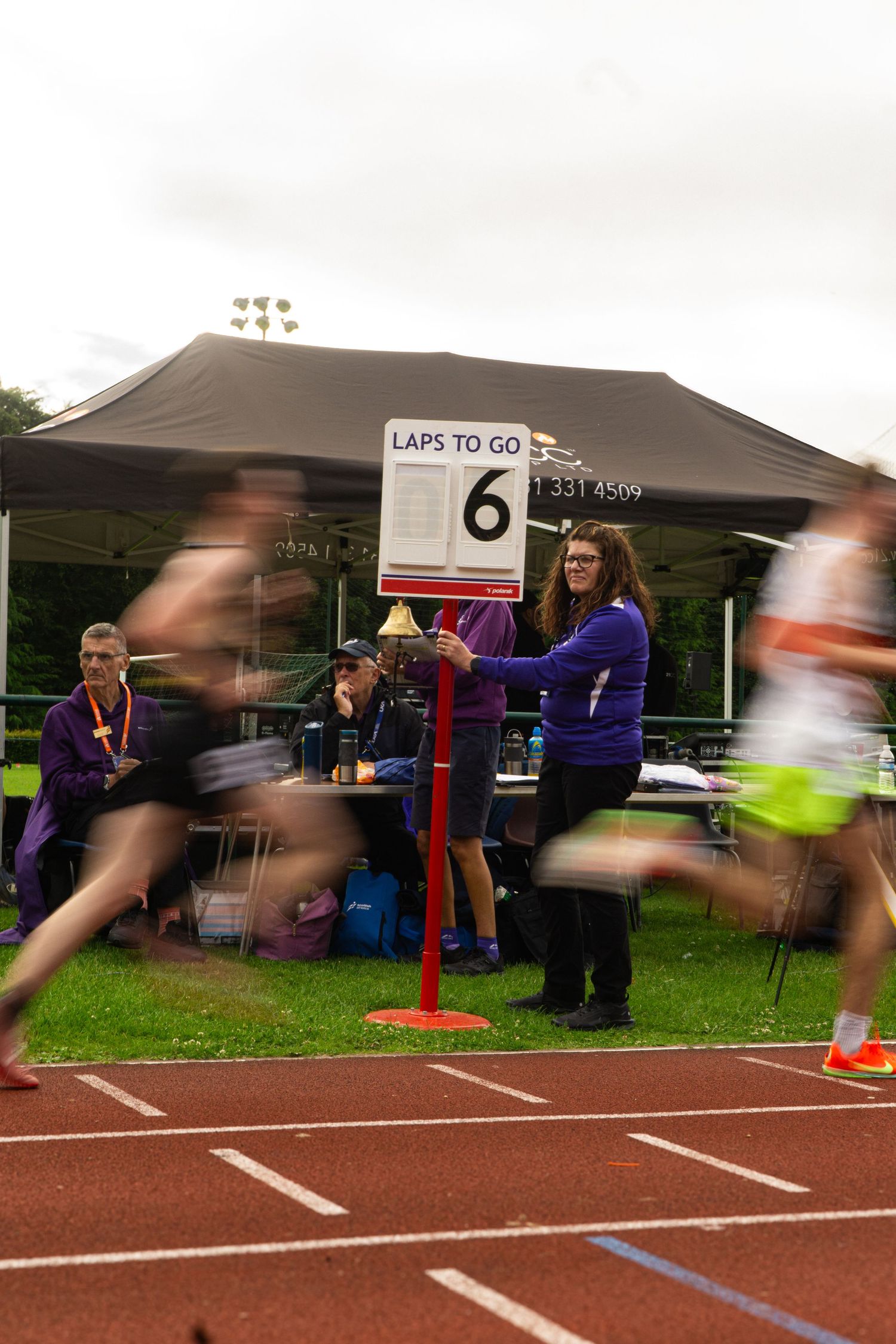 Track race with motion-blurred runners passing a lap counter sign showing 6 laps to go at an outdoor athletics event.