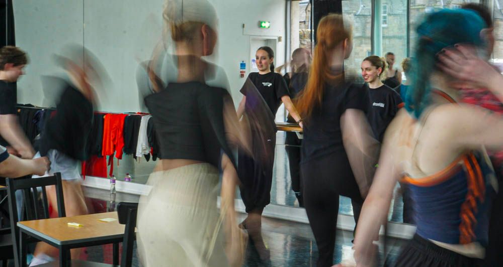 Motion-blurred dancers moving energetically in a bright dance studio with mirrors and chairs.