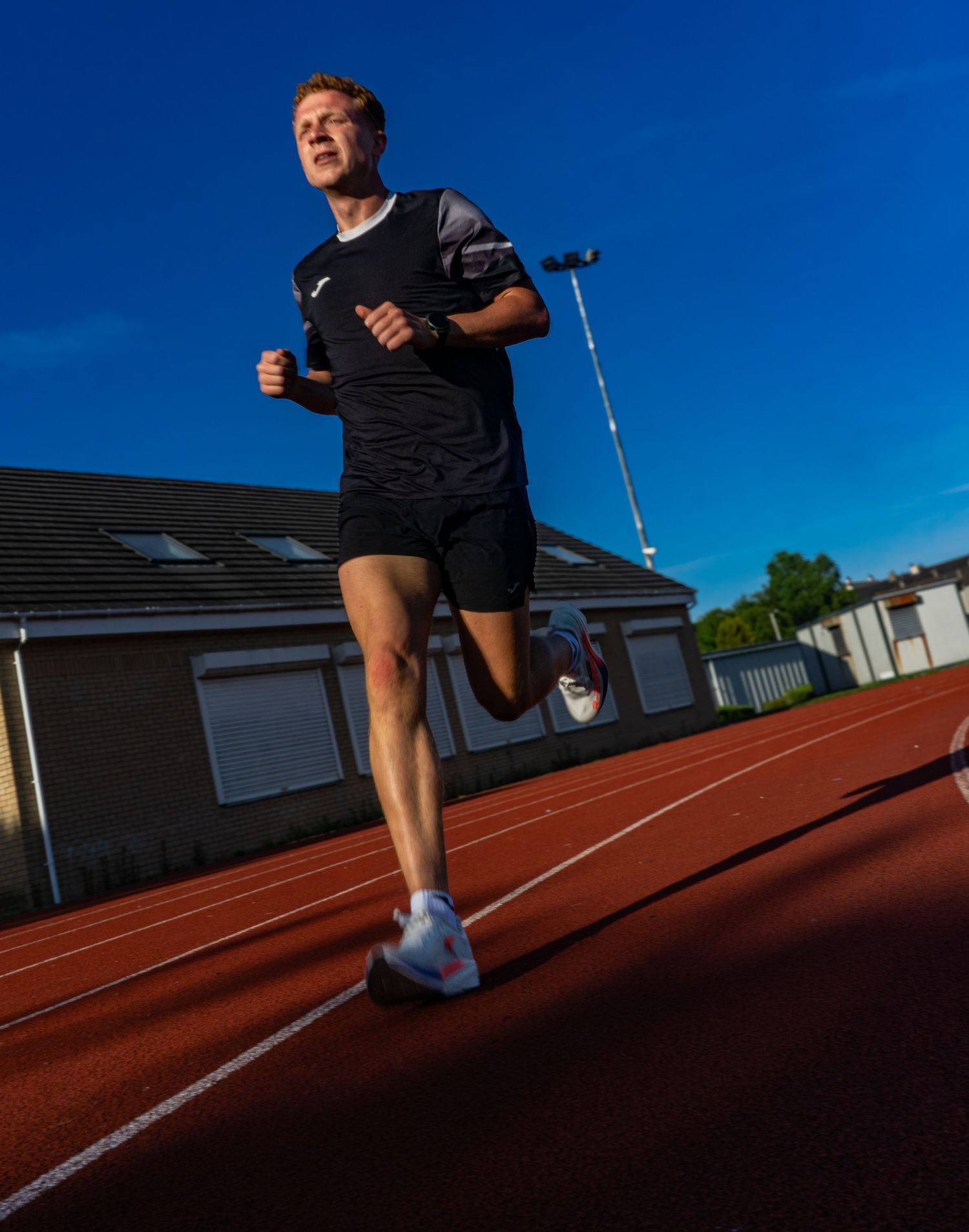 Male runner sprinting on a red athletics track wearing black shorts and white shoes under a vivid blue sky.