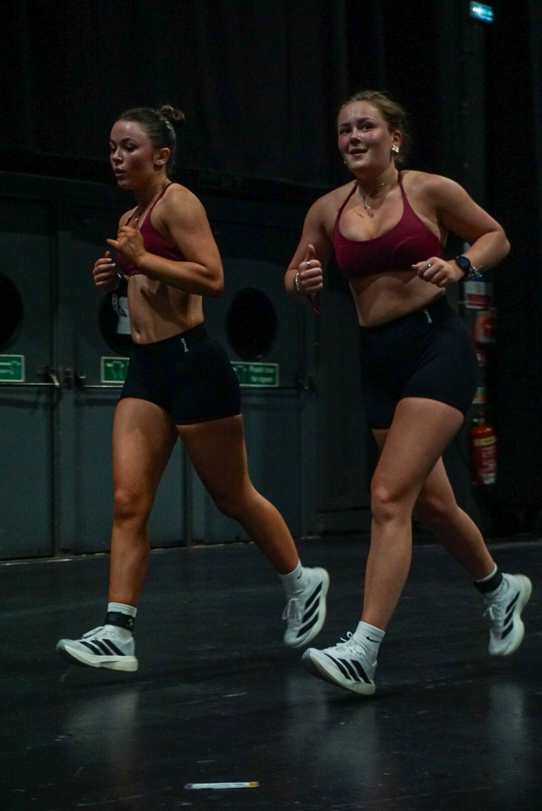 Two athletic women in matching red sports bras and black shorts jogging together during a fitness competition.