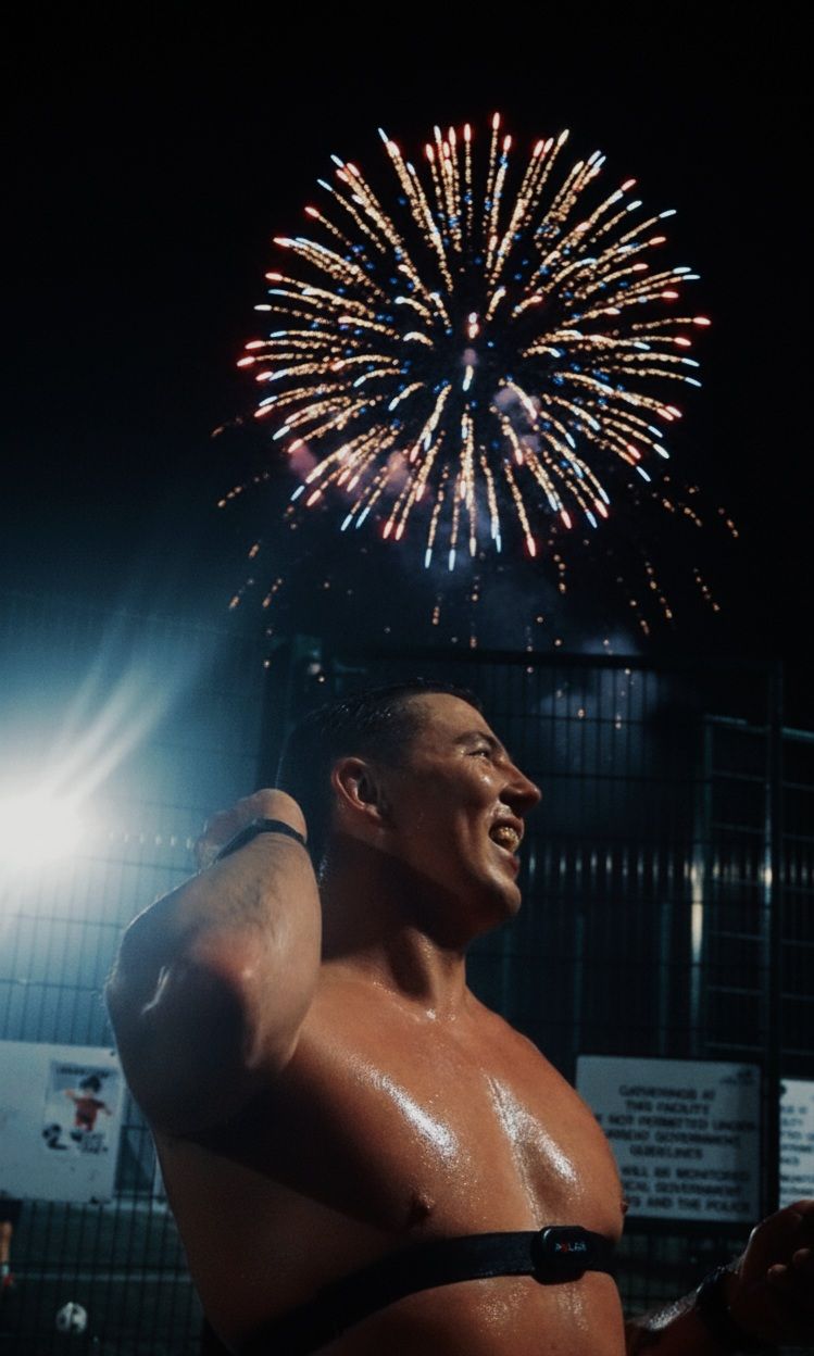 Muscular shirtless athlete wearing a heart rate monitor gazes at colorful fireworks in the night sky.