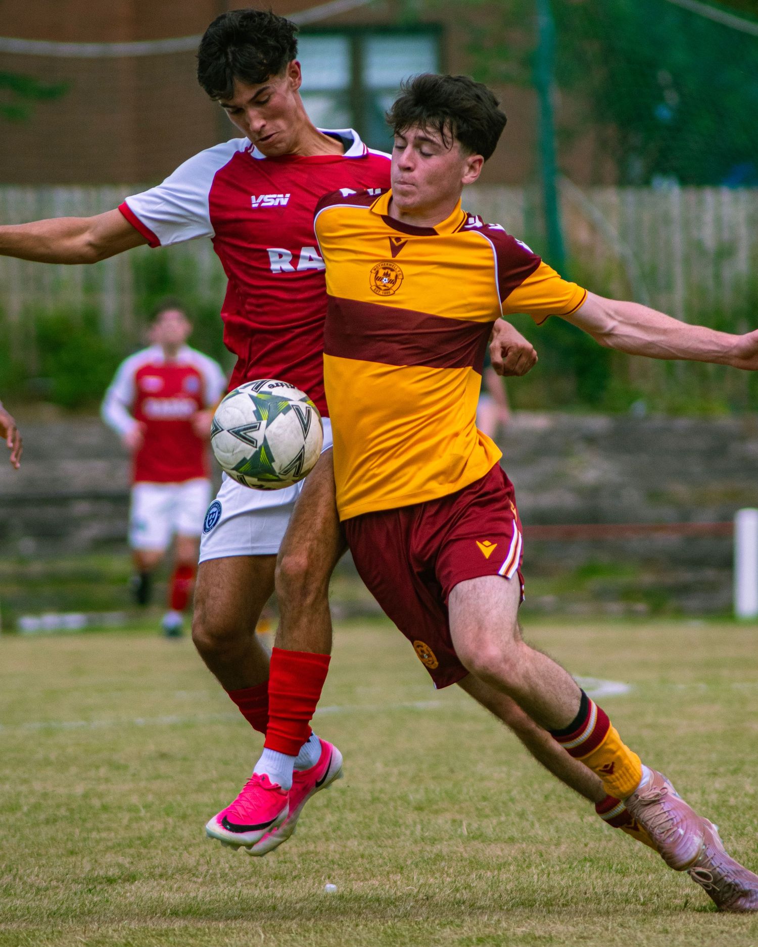 Two youth soccer players battle for the ball during a competitive match, wearing red-white and yellow-red kits.