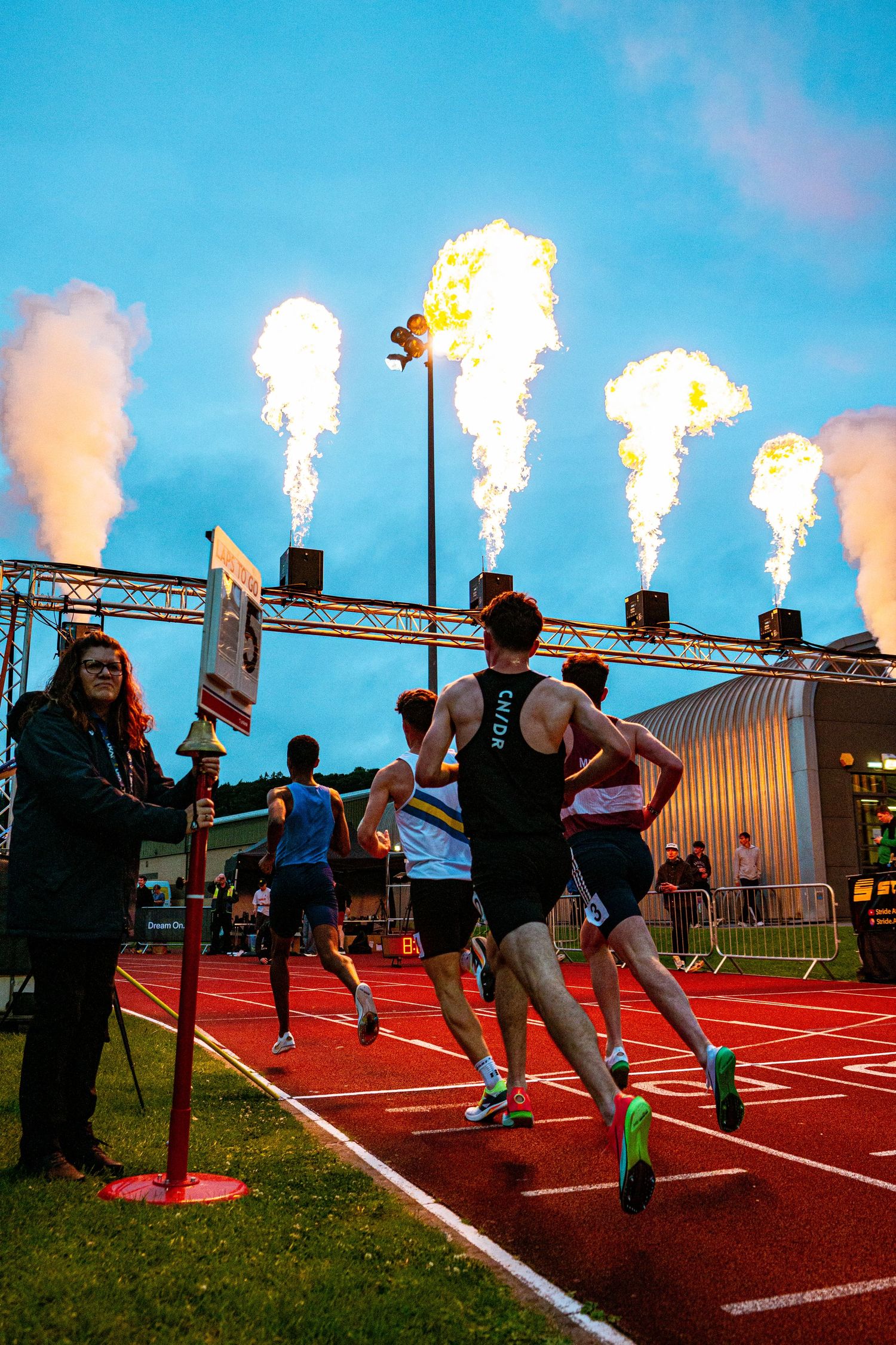 Athletes race on a red track at dusk with dramatic fire jets blazing overhead at an outdoor athletics event.