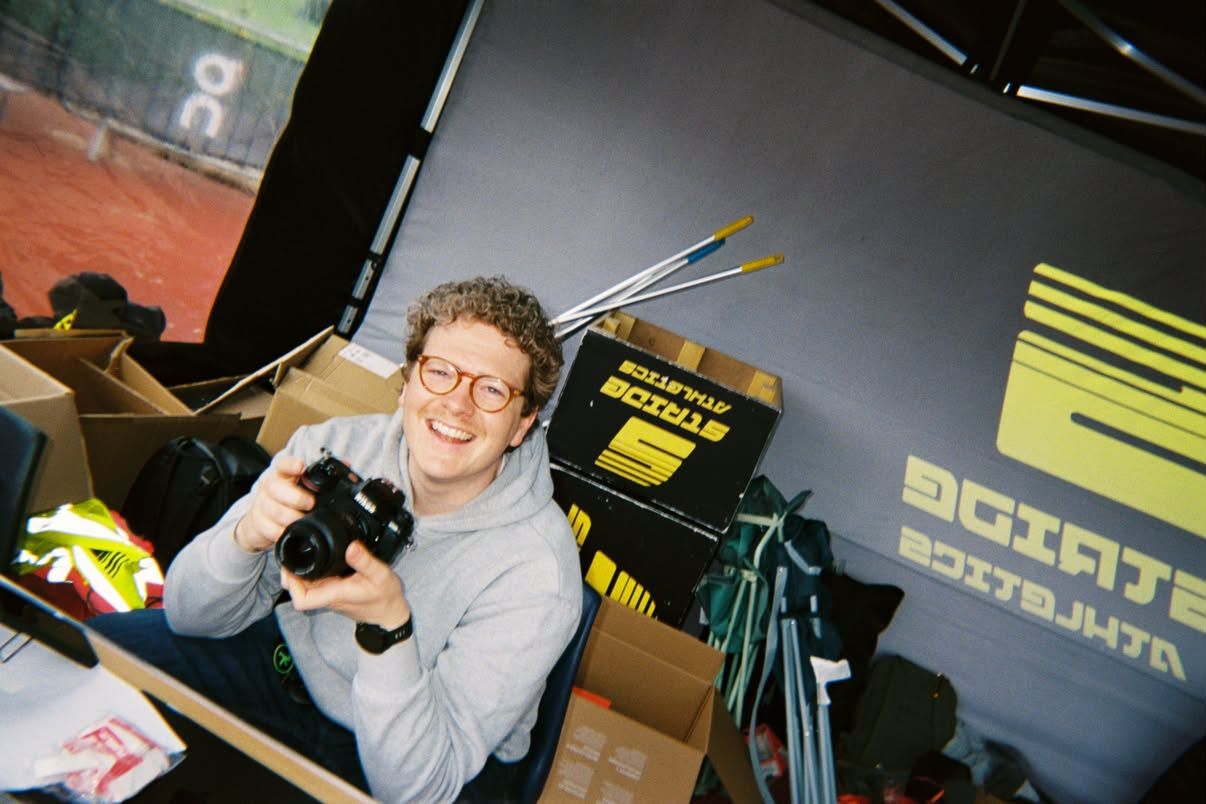 Smiling photographer holding a camera indoors near tennis courts, surrounded by boxes and sports equipment.