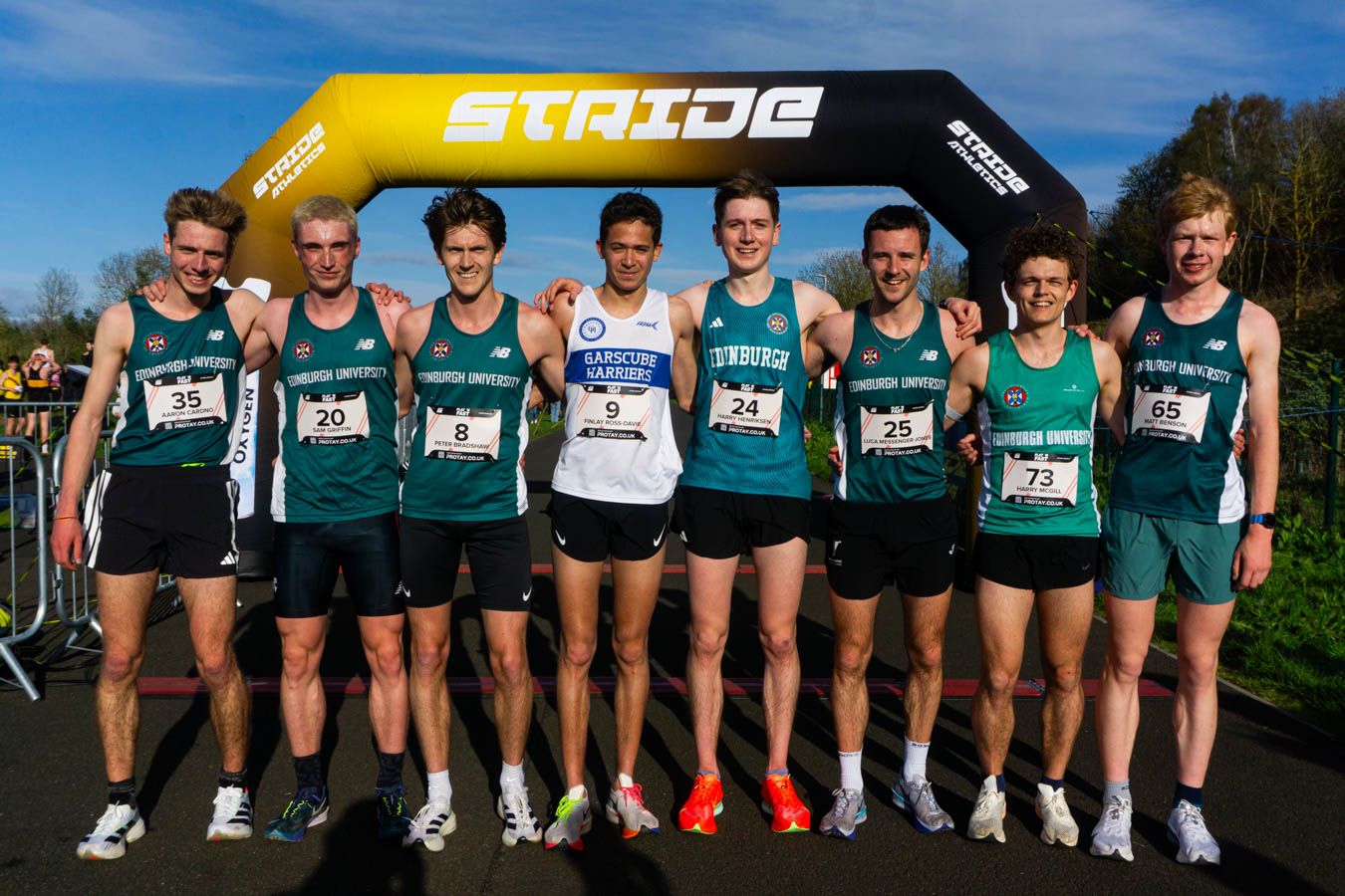 Group of eight male runners in green team uniforms posing together under a STRIDE finish arch.