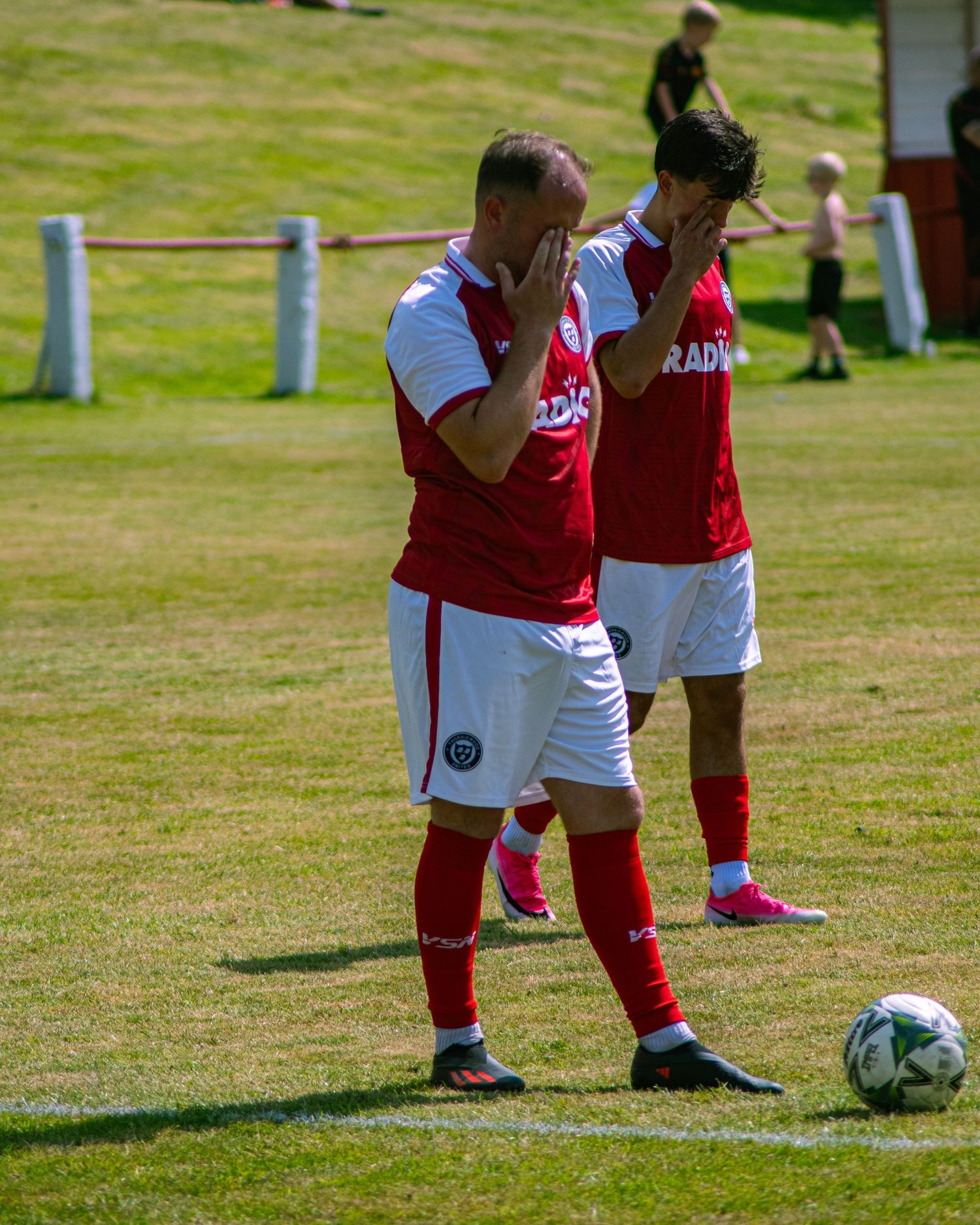Two footballers in red and white kits cover their faces on a sunny grass pitch during a match.