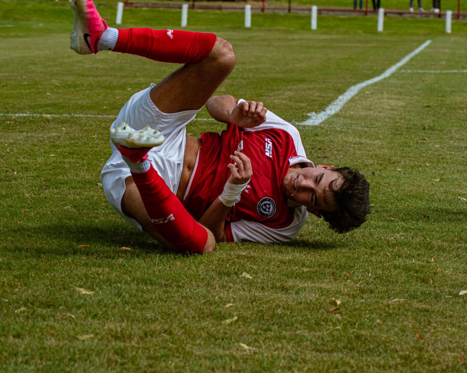 A footballer in red and white kit lies on the grass after a challenge.