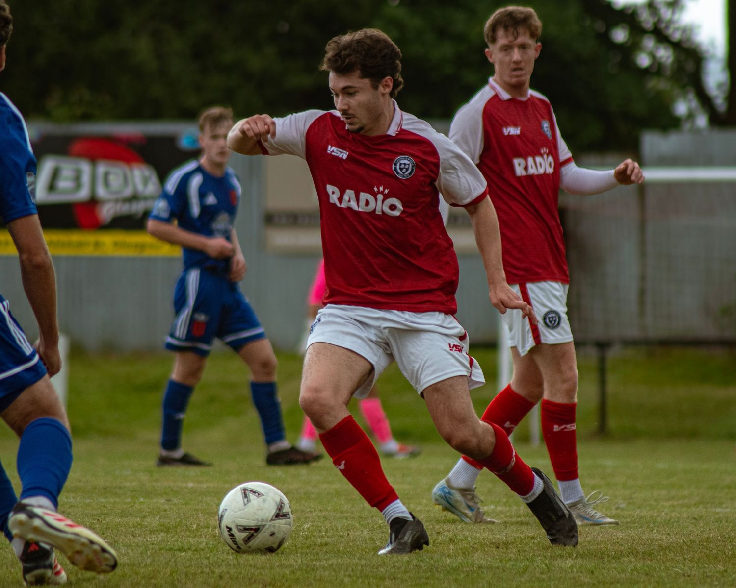 A young footballer in red and white Radio-sponsored kit dribbles the ball during a grassroots match.