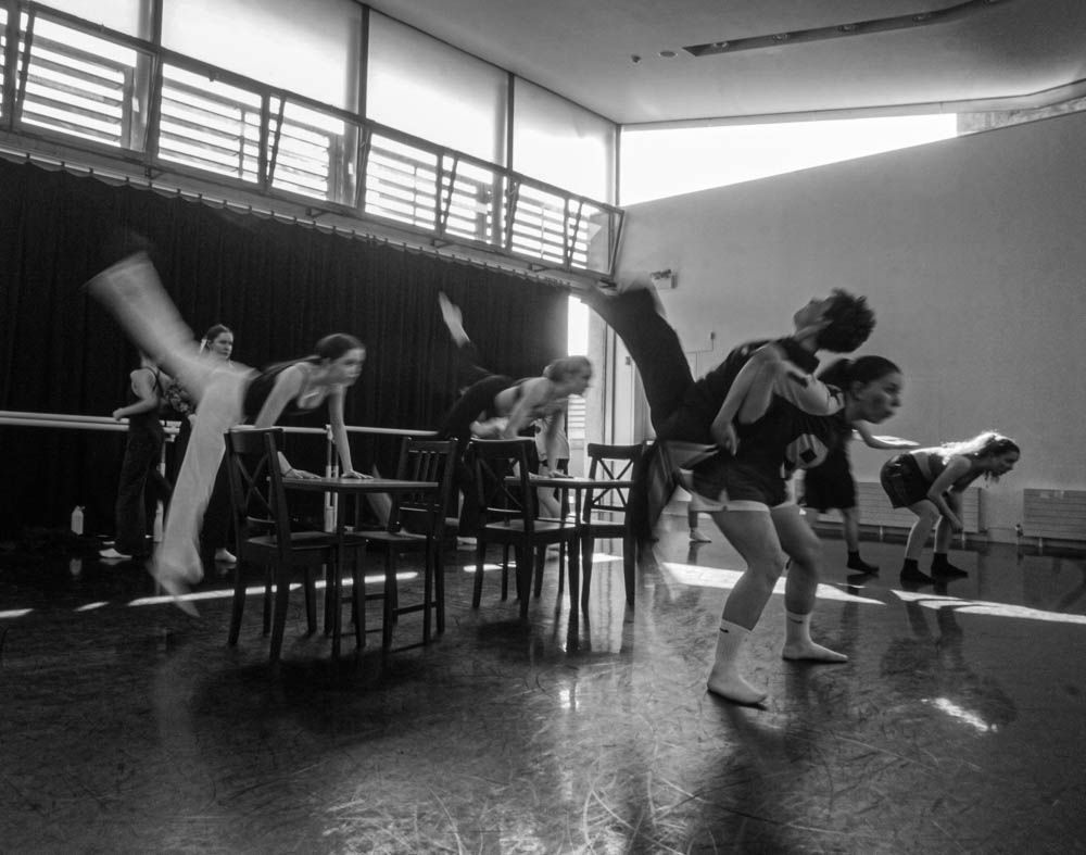 Black and white photo of ballet dancers practicing high kicks at the barre in a dance studio.