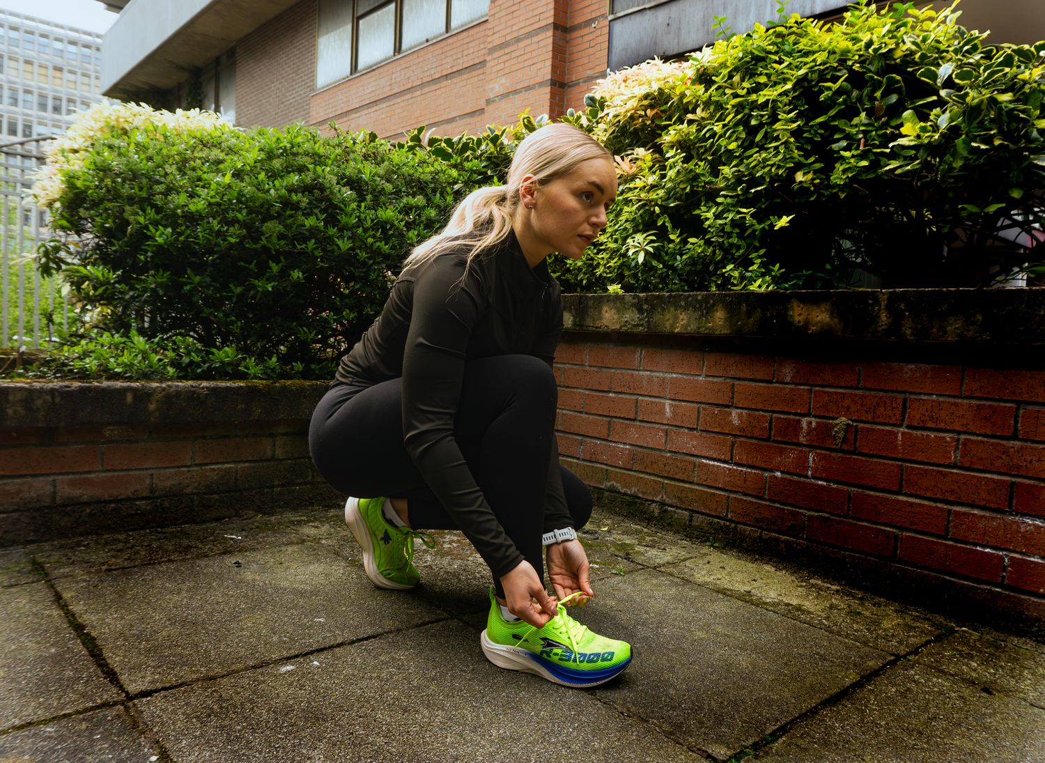 Woman in black athletic wear tying neon green running shoes outdoors on urban pavement.
