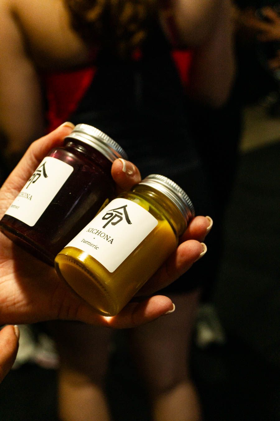 Hand holding two small jars of Michona honey, one dark and one golden, with silver lids.